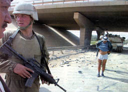 An elderly Iraqi man stopped by US soldiers before he is allowed to pass under a bridge in Baghdad. He had to lift up his clothes to show that he doesn't have explosives around his waist. (Alquds, 4/11/03).